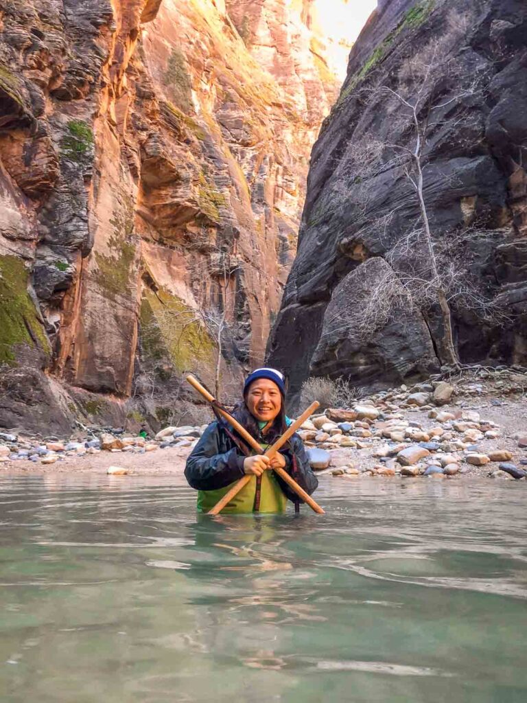 cat xu wading through the narrows holding some wlaking sticks with an x in front of her body