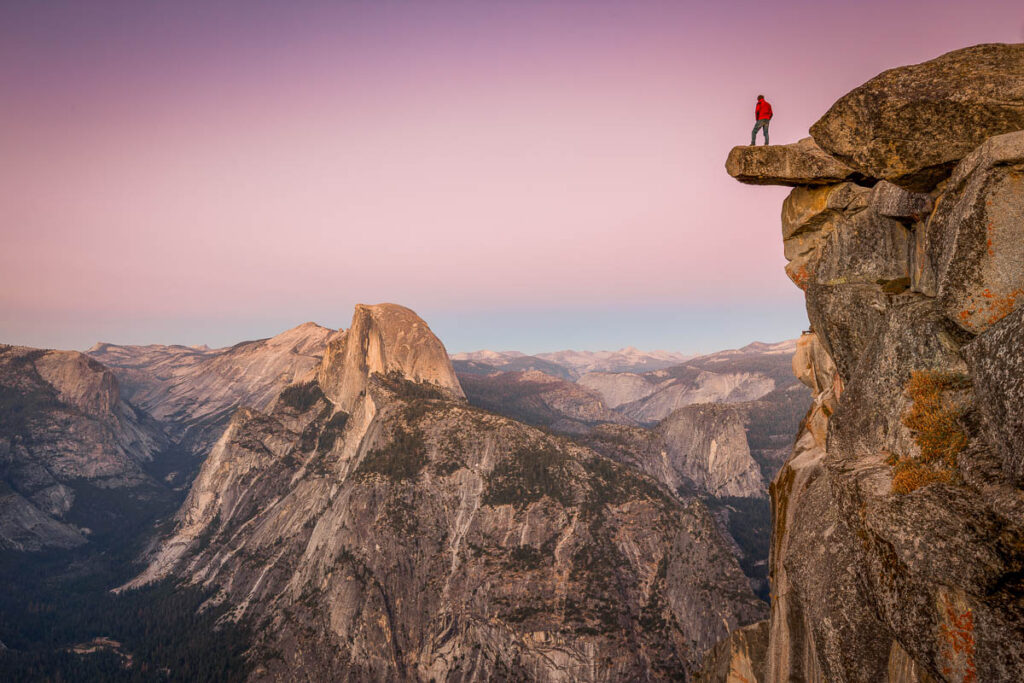 A fearless male hiker is standing on an overhanging rock at Glacier Point enjoying the breathtaking view towards famous Half Dome in beautiful post sunset twilight in summer, Yosemite National Park, California