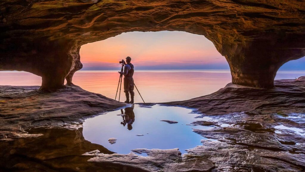 A photographer with tripod, standing in the mouth of a Michigan sea cave, is silhouetted by a colorful sunset over Lake Superior.