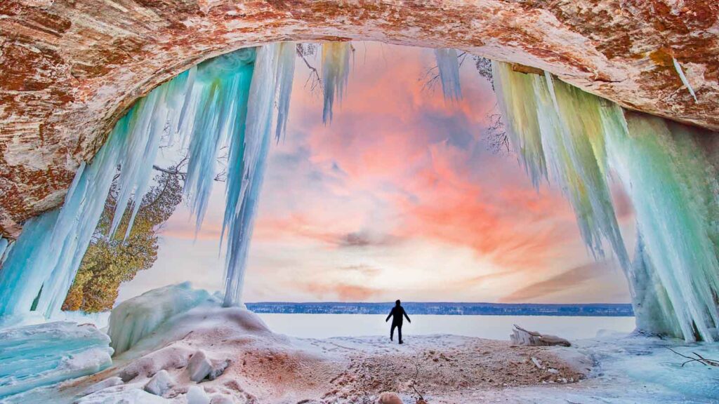 Human figure silhouette at entrance to stunning ice cave during winter sunrise with blue and green icicles