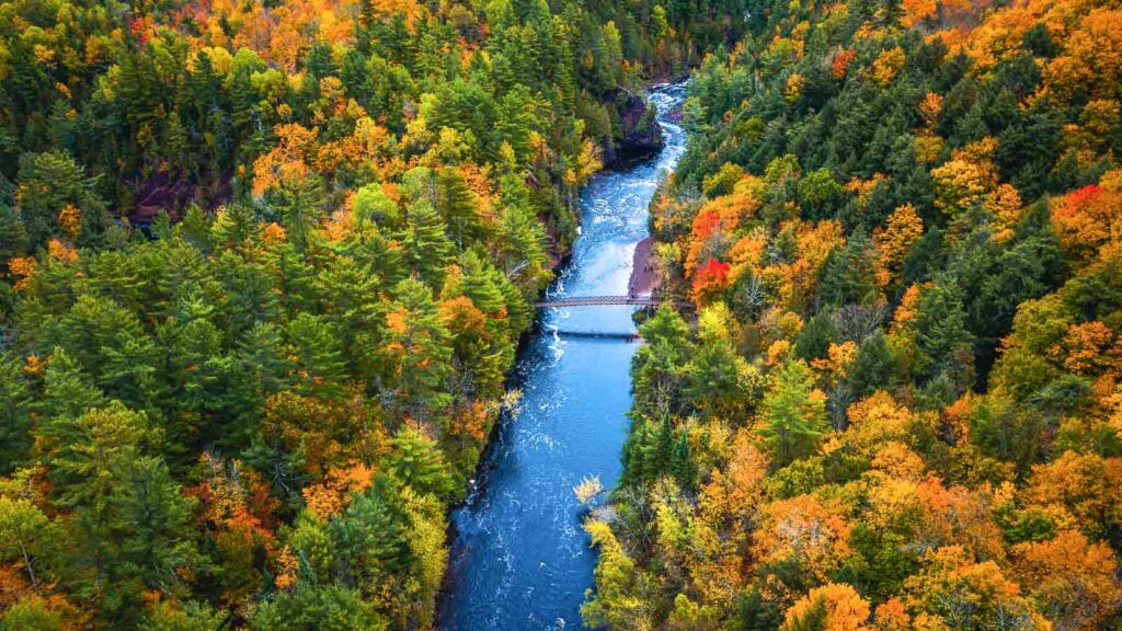 Beautiful travel aerial of a pedestrian foot bridge crossing the bright blue water of the Bad River at Copper Falls with colorful fall foliage lining the river banks in autumn in Mellen, Wisconsin.