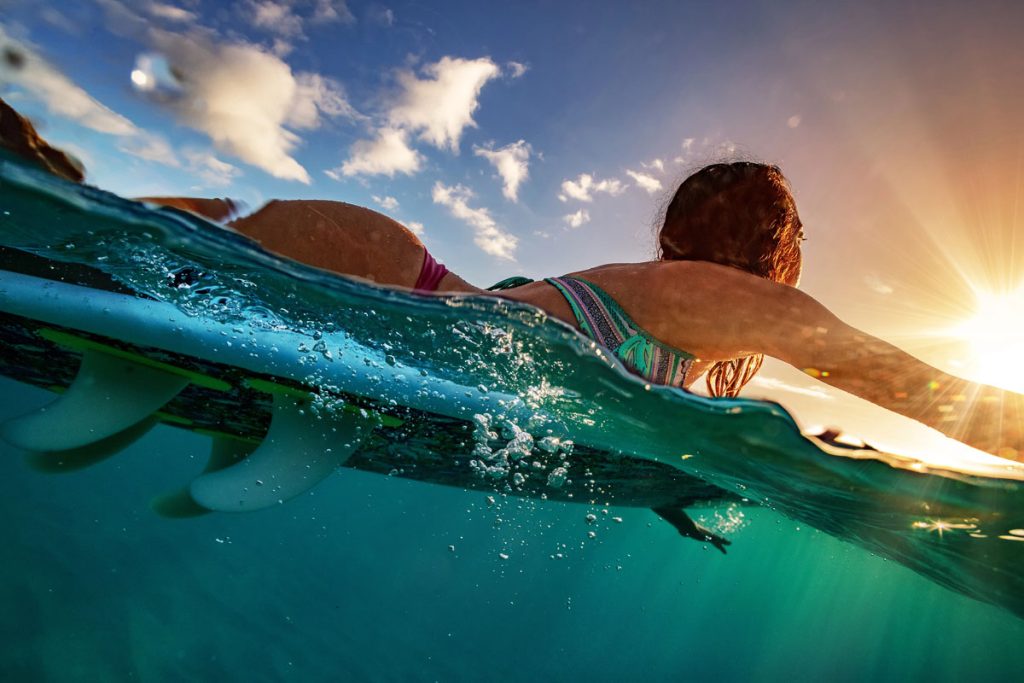 Half water shot of a surfer girl paddling on a board to the sun