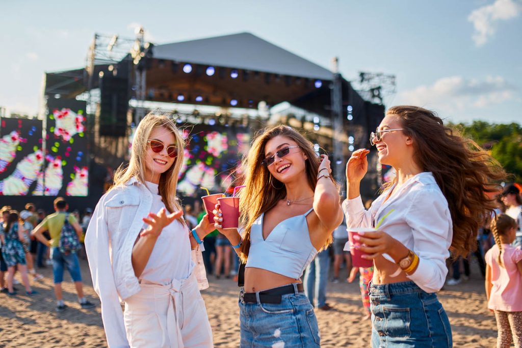Smiling female friends with sunglasses hold cold drinks, dance near stage in sunny day. Group of girls enjoy summer music festival on sandy beach. Party, fun, youth lifestyle at sea shore event.