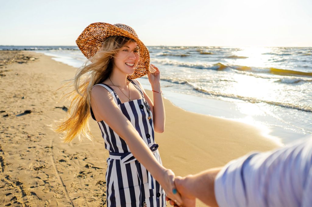 Smiling woman holding boyfriend’s hand while walking on sandy shore
