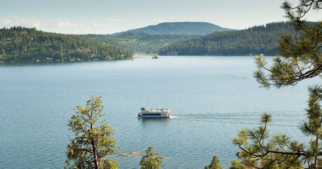 A tour boat cruises around Lake Coeur d'Alene, Idaho on blue water with background mountains and framed by pine trees.