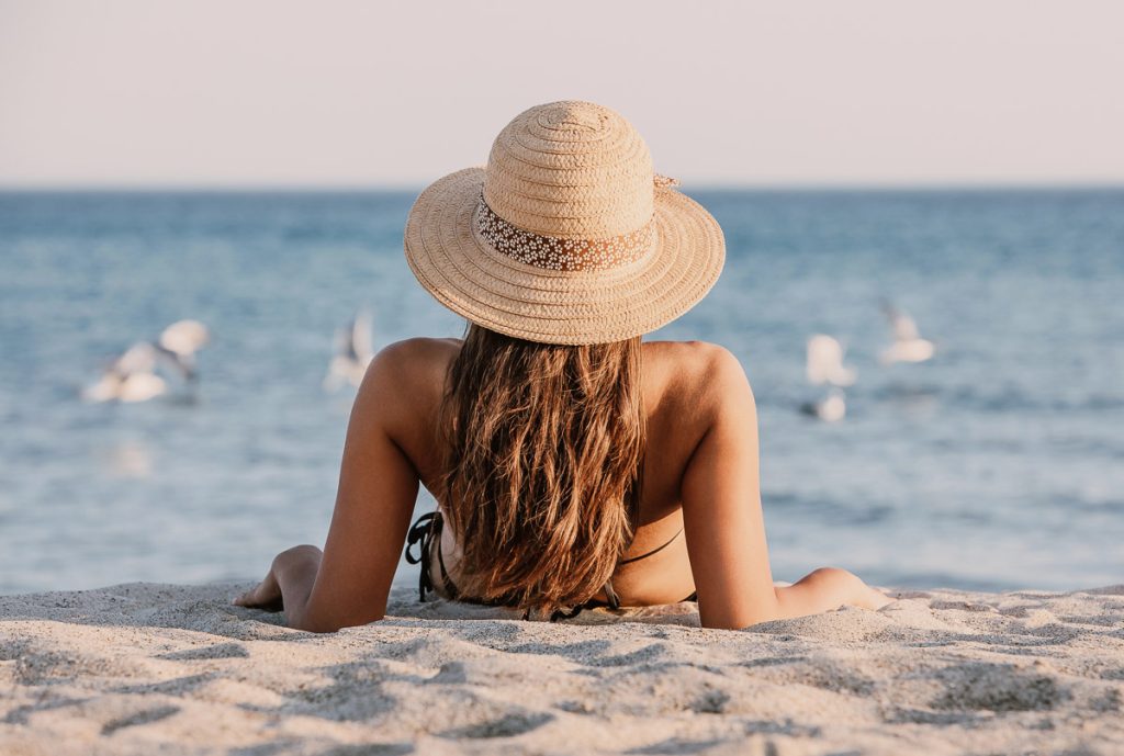 Young girl in a bathing suit and hat sitting on the sand on the beach, looking to the seagulls in the sea. Beautiful lady is facing the ocean with her back to the lens