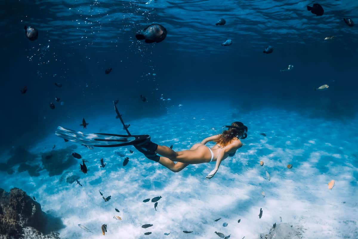 Free diver woman with fins glides over sandy bottom with fishes in transparent ocean